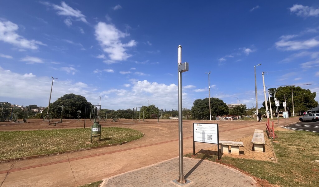 Uma área pública aberta em um parque, sob céu azul com algumas nuvens. No centro há um poste metálico alto e, ao lado, uma placa informativa próxima a um banco de concreto. Ao fundo, vê-se um espaço amplo de terra batida com equipamentos de academia ao ar livre. Há algumas árvores grandes e postes de iluminação espalhados pelo local. À direita, há uma calçada, pessoas caminhando e parte de um estacionamento com carros.